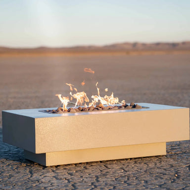 A white rectangular outdoor metal powdered coated fire pit with flames on top placed on a cracked, dry desert ground 