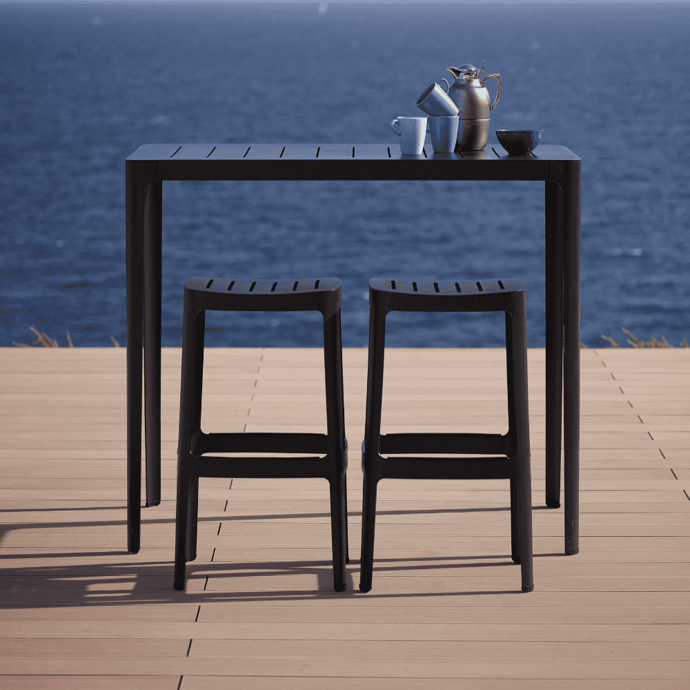Boxhill's Cut Outdoor Aluminum Bar Table Black lifestyle image with Cut High Outdoor Bar Chair on wooden platform. at seafront