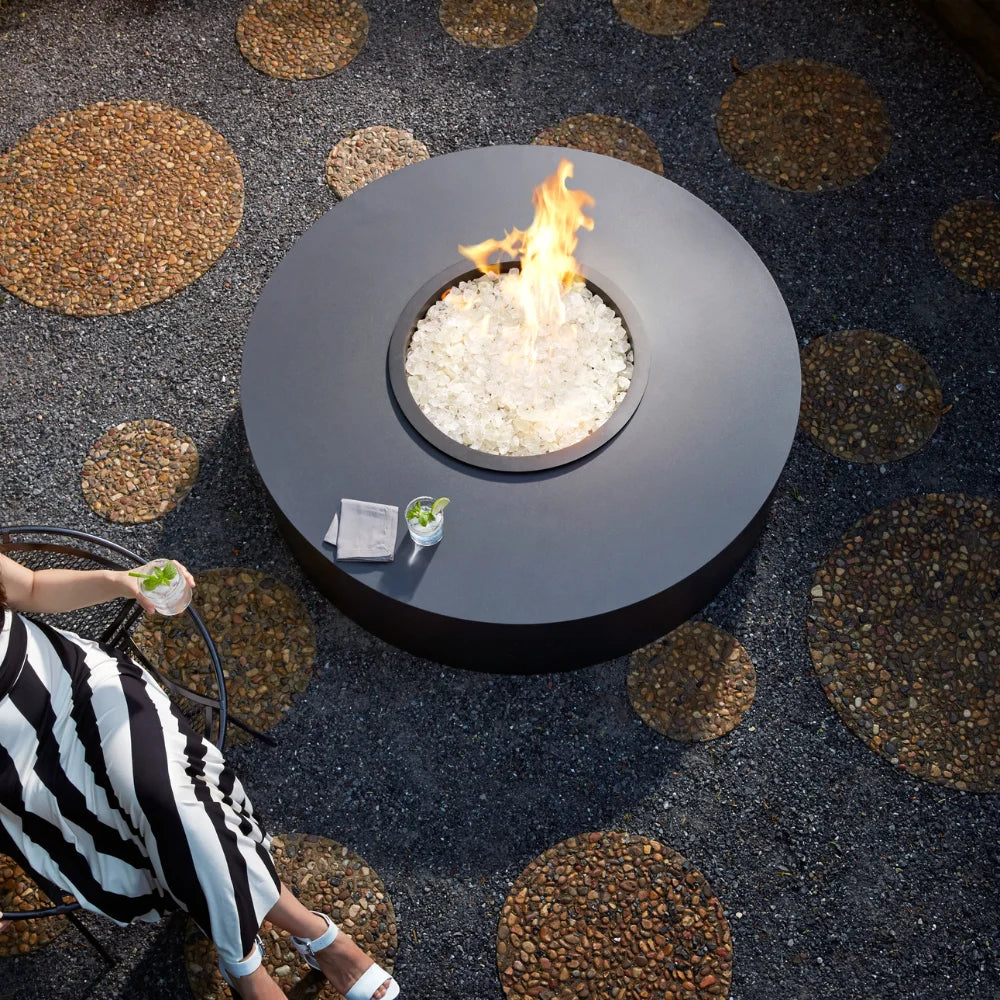 Modern black round fire pit table with white glass rocks and flames on a patterned stone patio