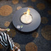 Modern black round fire pit table with white glass rocks and flames on a patterned stone patio