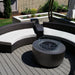 Patio seating with black round fire pit table with wind guard and curved black wicker sofa with white cushions on a wooden deck