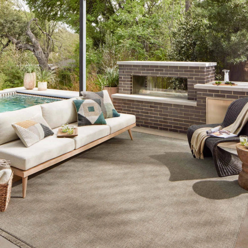A large textured neutral outdoor area rug in a modern patio setting, featuring a light wood sofa with patterned pillows, a brick fireplace, a pool, and a chair with a throw blanket.