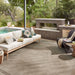 A large textured neutral outdoor area rug in a modern patio setting, featuring a light wood sofa with patterned pillows, a brick fireplace, a pool, and a chair with a throw blanket.