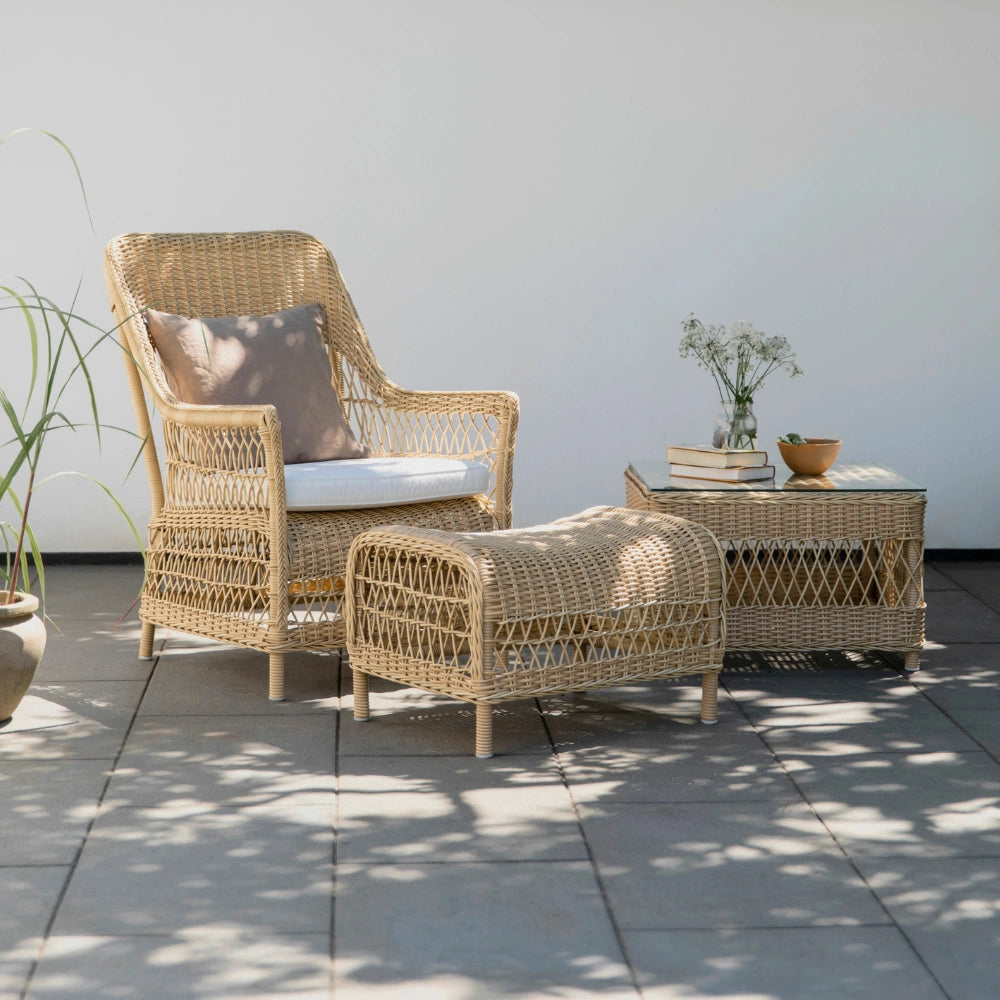 Natural wicker outdoor woven stool with matching armchair and side table featuring white cushions.
