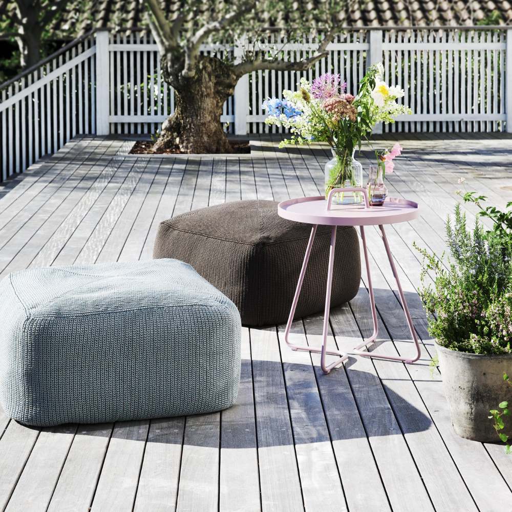 Boxhill's Divine Fabric Outdoor Footstool lifestyle image on wooden platform with pink round side table and flowers on top of it