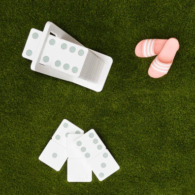 A set of white dominoes with pale blue-gray pips and a small pile of pink-and-white striped dominoes, arranged on green artificial grass.