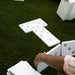 White outdoor domino tiles with pale blue-green pips arranged on green artificial turf, with a person's hand placing a tile in the foreground.