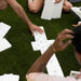 Several people sitting on artificial green grass play with white domino tiles arranged in a cross shape.