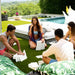 Four people sitting on a luxurious outdoor patio with a pool, lawn, and lounge chairs, playing with white foam blocks. The scene is bright and sunny with lush green landscaping in the background.