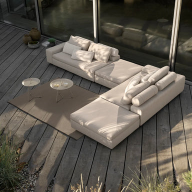 L-shaped light beige outdoor sectional sofa against glass walls on a wooden deck patio, with two round tray tables in front