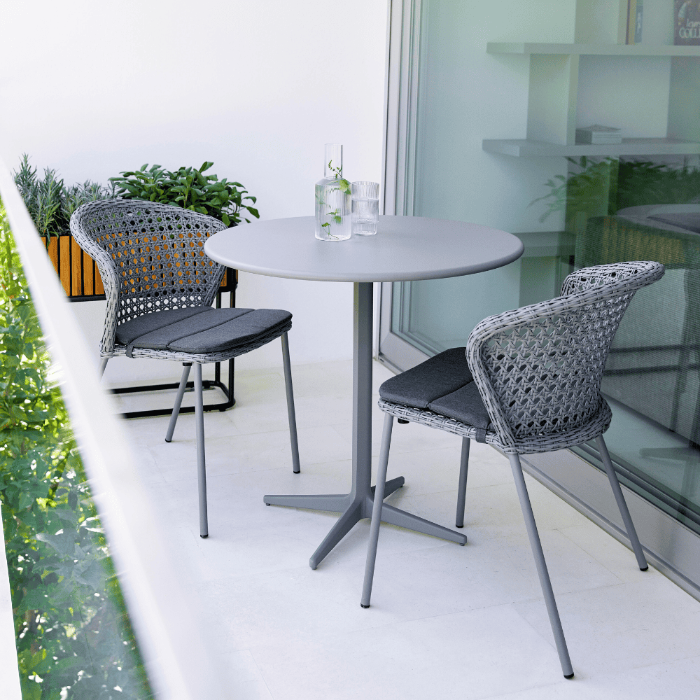 Boxhill's Drop Round Outdoor Cafe Table Light Grey lifestyle image on balcony beside sliding glass wall with 2 glass of water and water container on top, and 2 dining chairs and plants at the side