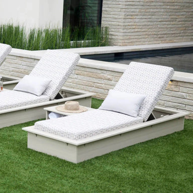 Two outdoor chaise lounge chairs with patterned white cushions and light gray frames, placed on green grass beside a stone wall and pool.