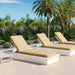 Two white outdoor chaise lounge chairs with beige cushions are arranged on a patio with palm trees and green plants in the background.