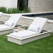 Two outdoor chaise lounge chairs with patterned white cushions and light gray frames, placed on green grass beside a stone wall and pool.