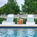 Two outdoor lounge chairs with pale blue cushions sit on white bases beside a swimming pool, with a small carved wooden side table between them and trees in the background.