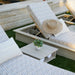 Outdoor patio lounge chaise with light gray and white geometric patterned cushions, a small wooden finish outdoor side table with a potted plant, and a straw hat resting on one chair, set on green grass.