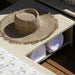 A woven straw sun hat with a wide frayed brim rests on a light wood shelf, with two rolled white towels underneath in a sunlit setting.