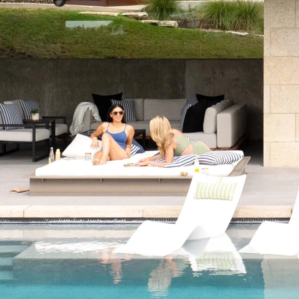 Two women relax on white lounge chairs by a swimming pool in a modern outdoor patio setting with a sofa, striped pillows, and a grassy upper level in the background.
