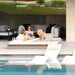 Two women relax on white lounge chairs by a swimming pool in a modern outdoor patio setting with a sofa, striped pillows, and a grassy upper level in the background.