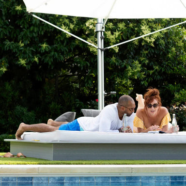 A man and a woman relax on a white outdoor daybed beside a swimming pool, with a large white patio umbrella and lush green foliage in the background.