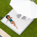A woman in a green swimsuit reclines on a large white outdoor daybed with striped pillows, under a white patio umbrella on green grass.