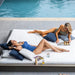 Two women relax on white lounge chairs by a swimming pool, wearing blue and olive green swimwear, with pillows, magazines, and a bottle on the chairs.