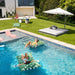Two women relax on tropical green palm-leaf inflatable pool float loungers in a swimming pool, with a modern house, green lawn, patio daybed, and white patio umbrella in the background.