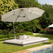 A large outdoor patio daybed with a matching cantilever umbrella in a lush green backyard next to a swimming pool. The daybed has a light beige cushion on a gray frame, with two accent pillows, and the umbrella is a light beige color with a silver metal pole.