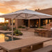 A large white cantilevered patio umbrella over a stone poolside deck at sunset, with a stucco house, dining chairs, and desert landscaping in the background.