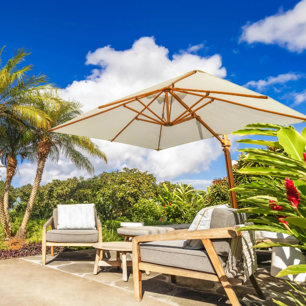 Outdoor patio furniture set under a large cream patio umbrella, featuring a wooden-framed lounge chair, a matching armchair, and a small side table, with palm trees and tropical greenery in the background under a blue sky.