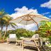 Outdoor patio furniture set under a large cream patio umbrella, featuring a wooden-framed lounge chair, a matching armchair, and a small side table, with palm trees and tropical greenery in the background under a blue sky.