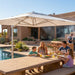 A large cream patio umbrella in use at a poolside outdoor dining area, with a family seated at a stone table under its shade. A stucco house, plants, and a cook in the background complete the lifestyle scene.