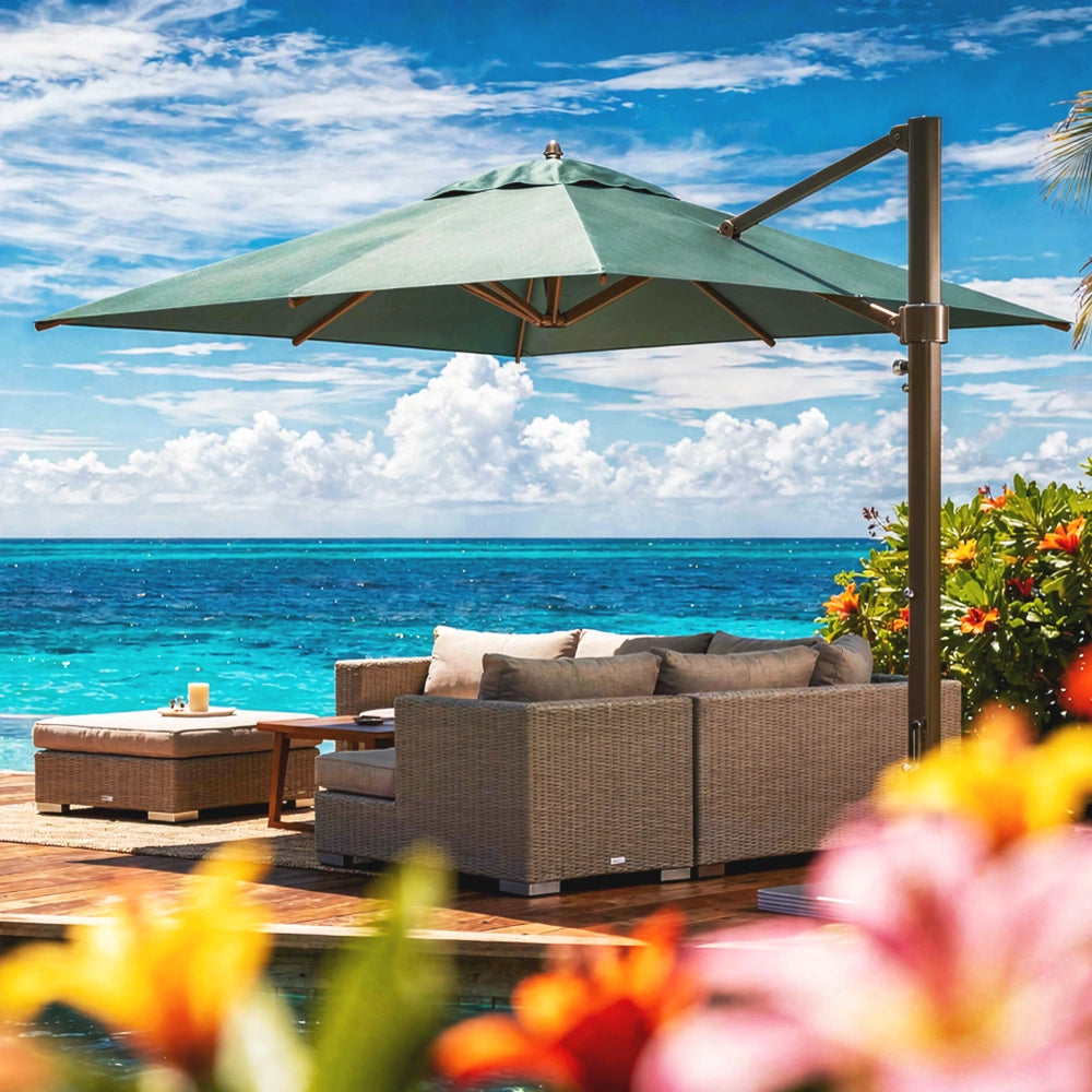 A large green cantilever patio umbrella over a wicker outdoor sofa set with cushions, next to a matching ottoman, on a wooden deck overlooking a bright blue ocean and sky.