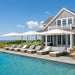 A row of white patio umbrellas and wicker lounge chairs with cushions arranged along a swimming pool beside a large coastal house under a bright blue sky.