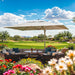 A large cream patio umbrella shading a wicker outdoor lounge set with cushions, set on a patio overlooking a golf course under a bright blue sky with clouds.