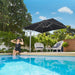 A woman in a black swimsuit sits at the edge of a swimming pool under a large black patio umbrella, with two white lounge chairs and green trees in the background.