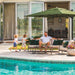 A man relaxes on a patio sofa beside a swimming pool, with two children running nearby under a large green patio umbrella. The setting is a modern home with glass doors and outdoor furniture.