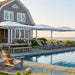 Outdoor poolside patio scene with a large gray shingled house, white patio umbrellas, striped lounge chairs, a dining table, and ocean view in the background.