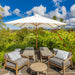 Outdoor patio lounge setup with a large white beach umbrella, four wooden lounge chairs with gray cushions, and a round coffee table, set against lush tropical greenery and a bright blue sky.