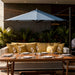 Outdoor patio dining setup with a large blue cantilever umbrella over a white wicker sofa with mustard yellow pillows, set with wine glasses and plates on a wooden table.