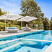 Two white outdoor patio umbrellas with matching round lounge chairs and blue striped pillows on a poolside deck, with a woman sitting on one chair and trees in the background.
