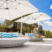 A woman sits on a round wicker daybed with blue and white striped pillows under a large white patio umbrella on an outdoor terrace. Another matching daybed and a second umbrella are visible in the background.