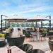 An outdoor patio dining area with multiple large cream umbrellas, woven black chairs, and tables on a wooden deck overlooking water and overwater bungalows under a blue sky.