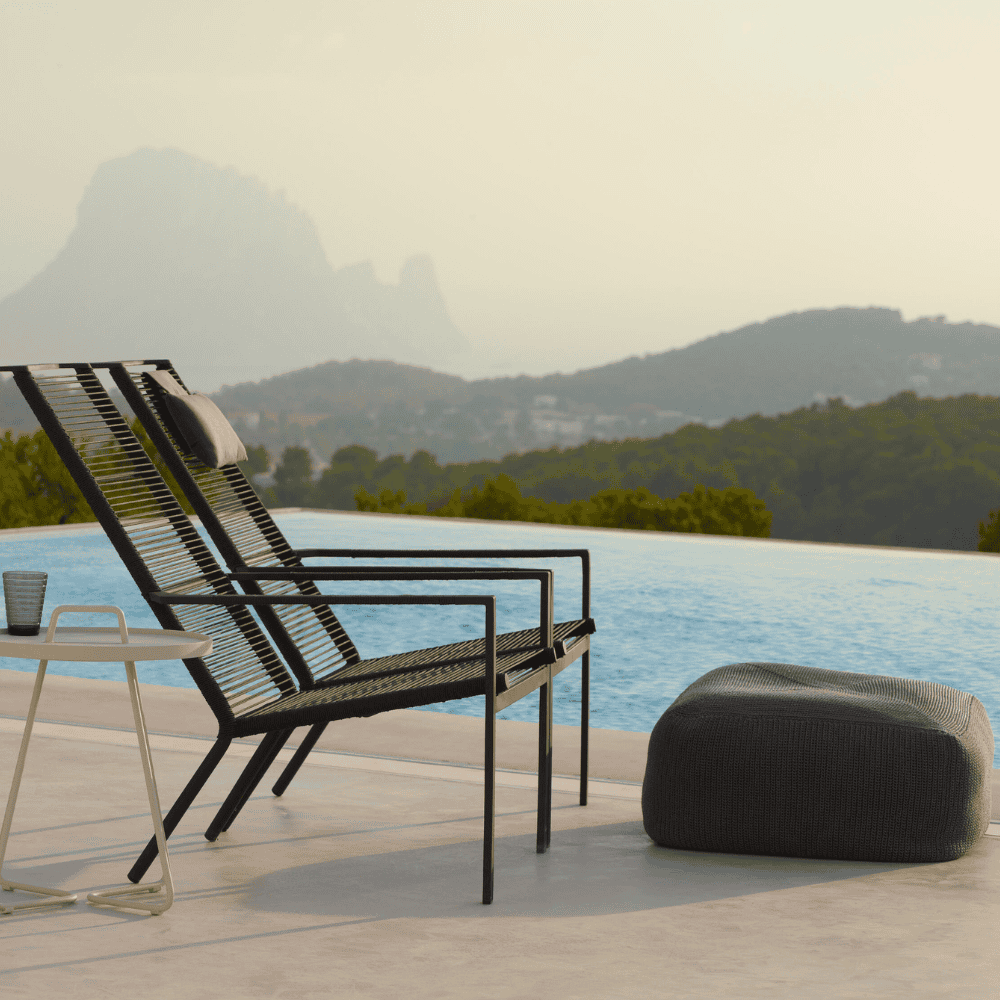 Boxhill's Edge Stackable Pool Side Highback Chair lifestyle image beside the pool with Divine Fabric Outdoor Footstool and round side table with glass of water on top