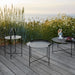 Three modern black metal outdoor tray tables in different sizes, placed on a wooden deck against tall grasses overlooking the sea