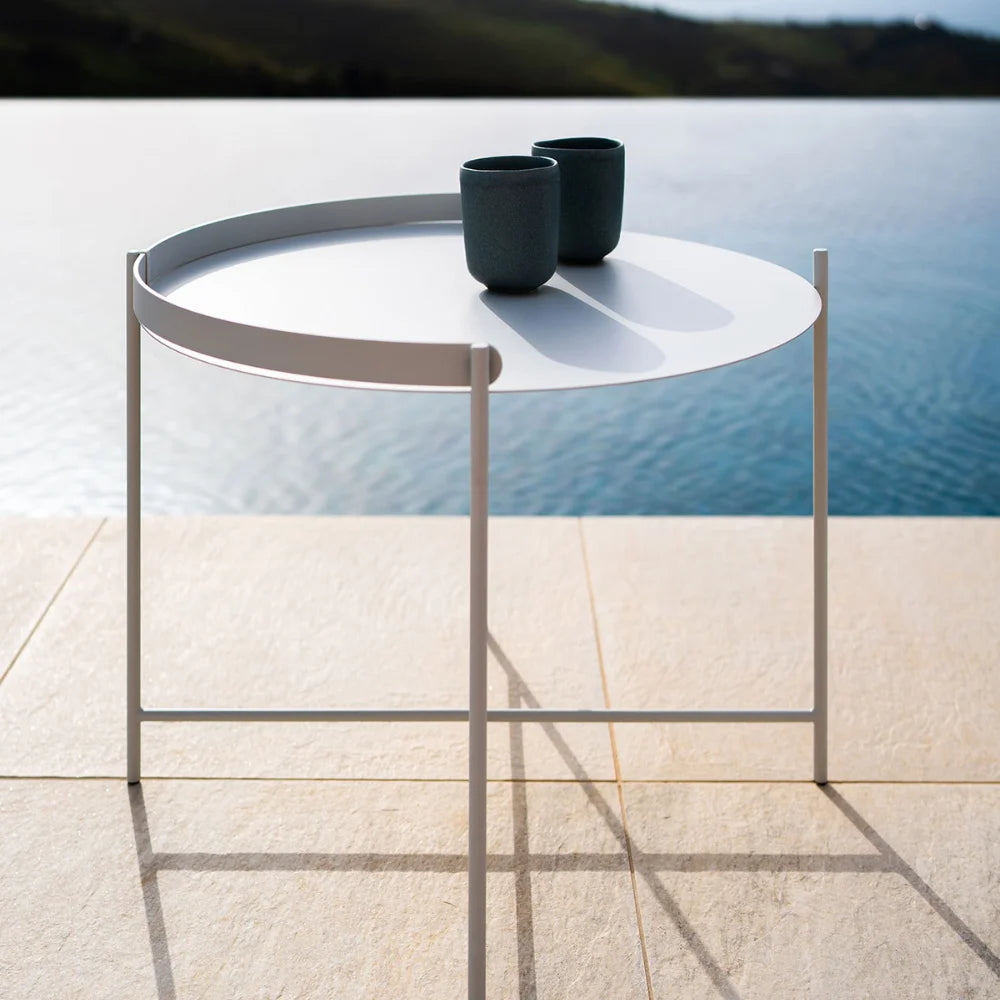 White metal round outdoor tray table with two black cups on top, placed on a tiled surface beside the pool