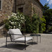 Outdoor lounge chair with white cushions and round metal outdoor tray table, placed on a patio near a stone house with greenery