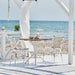 White rattan outdoor dining side chairs with a wooden pedestal table on a seaside patio.