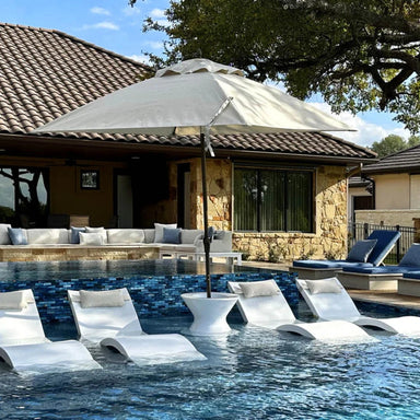 Large beige patio umbrella providing shade over partially submerged loungers in a shallow pool in front of a stone house.