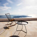 Black round metal outdoor accent side table and a rocking chair with a wooden seat, placed on a patio overlooking the sea and islands